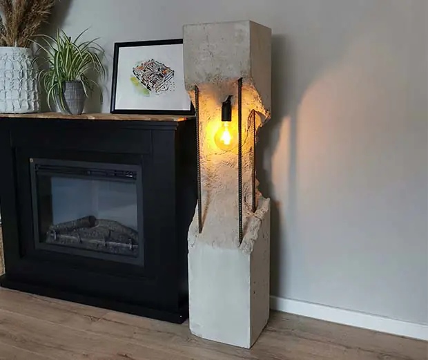 Industrielle Stehlampe aus Beton mit freiliegender Glühbirne neben einem schwarzen Kamin und Pflanzen.