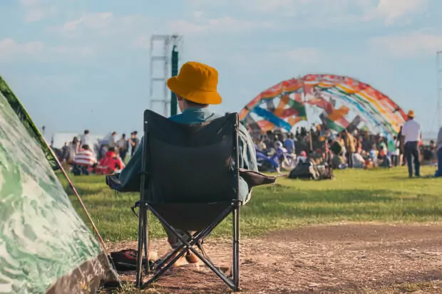 Auf einem Campingstuhl sitzt ein Mann mit orangenem Hut. Im Hintergrund sieht man das Treiben des Festivals, links neben dem Mann ein Zelt.