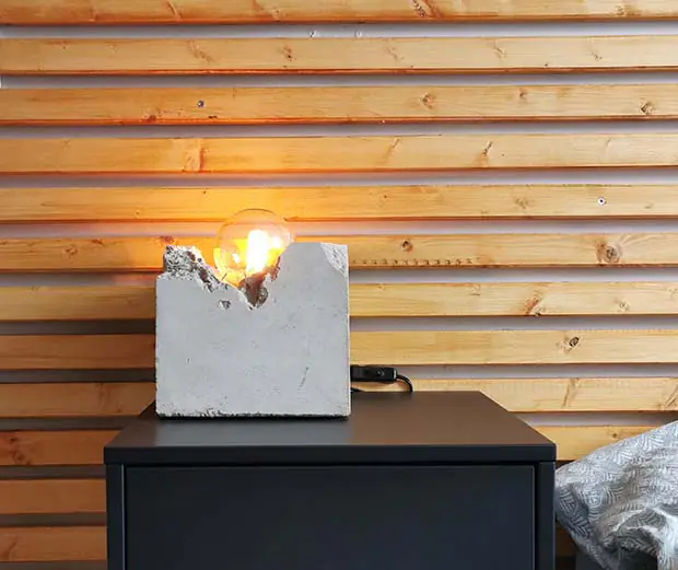 Moderne Tischlampe aus Beton mit freiliegender Glühbirne auf schwarzem Nachttisch vor einer Holzlattenwand.
