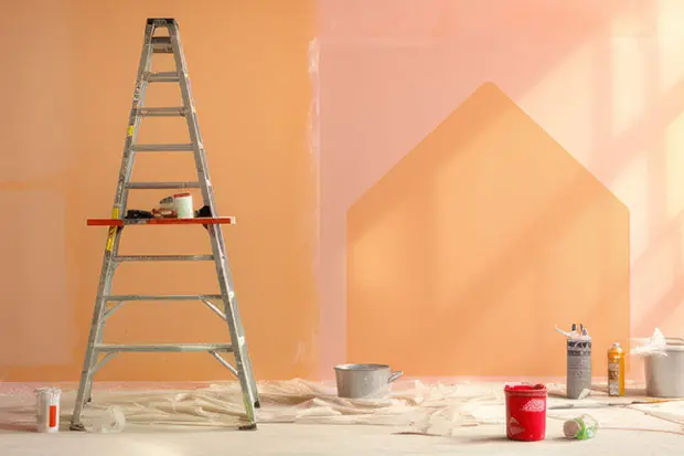 Leiter vor einer halb gestrichenen Wand in verschiedenen Orangetönen, auf dem Boden stehen Farbeimer und Malerutensilien, im rechten Bereich ist eine Hausform in einer anderen Farbe an die Wand gemalt.