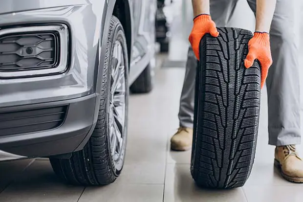 Mann mit orangefarbenen Handschuhen hält einen Autoreifen neben einem silbernen Fahrzeug in einer Werkstatt.