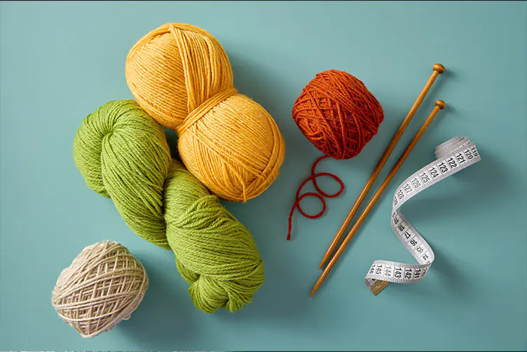Bunte Wollknäuel in Gelb, Grün, Rot und Beige mit Stricknadeln und Maßband auf türkisfarbenem Hintergrund.