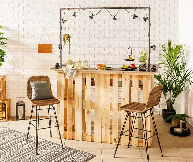 Die DIY-Palettenbar auf einer Terrasse. Vor ihr stehen 2 Barhocker. Sie ist dektoriert mit Lichterketten und Pflanzen.
