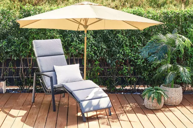Eine Sonnenliege steht auf der Holzterrasse vor einer grünen Hecke. Ein großer  Sonnenschirm spendet Schatten.