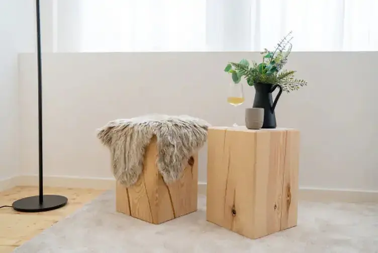 Zwei Holzblöcke als Hocker und Beistelltisch, einer mit Fellauflage, der andere mit Vase und Getränk, in einem hellen, minimalistischen Raum.