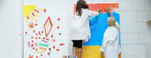 Zwei Kinder in weißen Kitteln bemalen eine weiße Wand bunt mit Pinseln und Händen; links Sonne und Regenbogen, rechts Flächen in Rot, Blau und Gelb.
