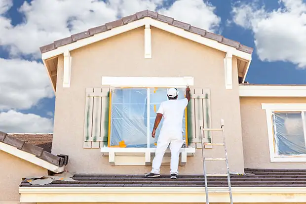 Ein Mann streicht eine Hausfassade in Beige bei schönem Wetter.