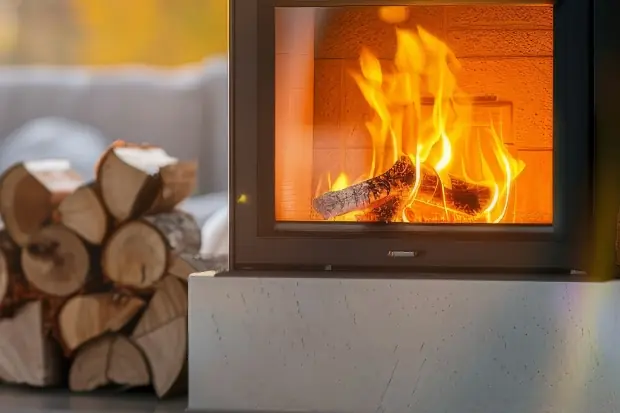 Moderner Kamin mit brennendem Feuer, daneben gestapeltes Kaminholz, im Hintergrund gemütliches Wohnzimmer mit Sofa und Decke – warme Lichtstimmung bei Sonnenuntergang