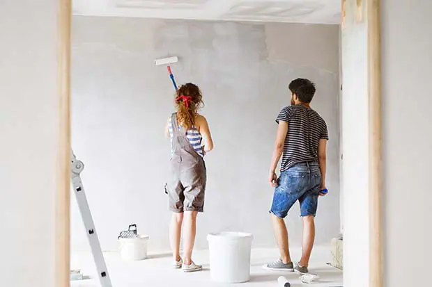 Junge Frau und junger Mann beim Streichen einer Wand in einem hellen Raum, die Frau benutzt eine Farbrolle, während der Mann zuschaut. Im Vordergrund steht eine weiße Farbeimer und eine Leiter.
